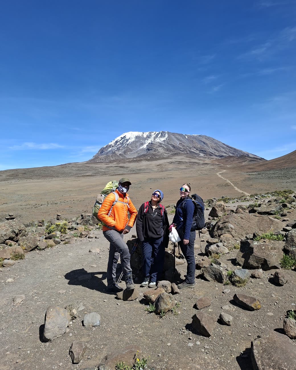 Three trekkers resting on the alpine-desert saddle on the Marangu route, the snow-streaked summit cone of Kibo rising behind them under a deep blue sky