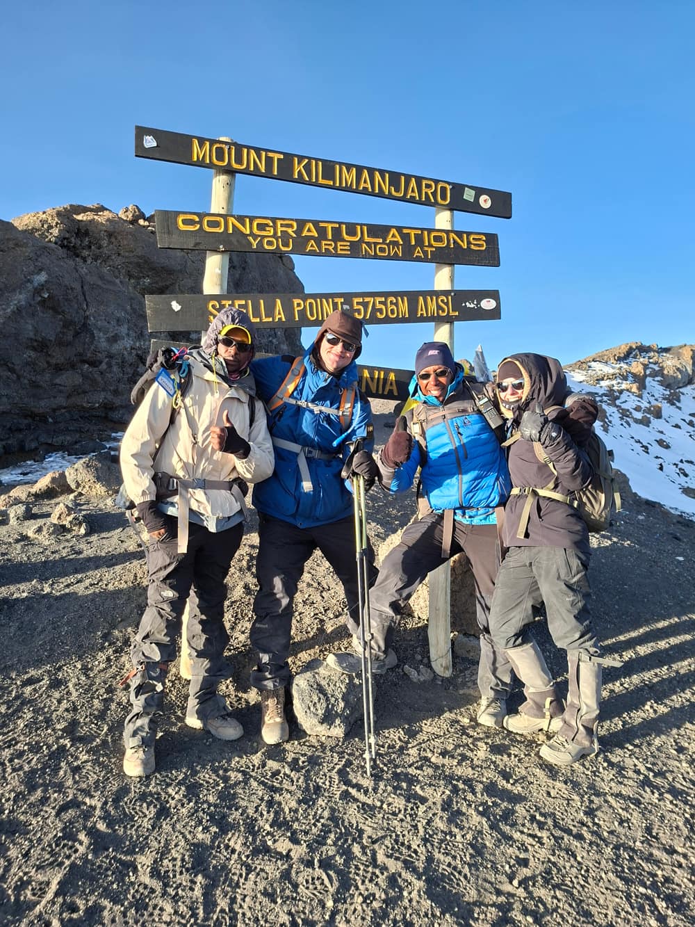 Four climbers in down jackets and gaiters standing at the carved Stella Point signboard reading 5,756 m, first sun on the rock and snow lying on the crater rim behind them
